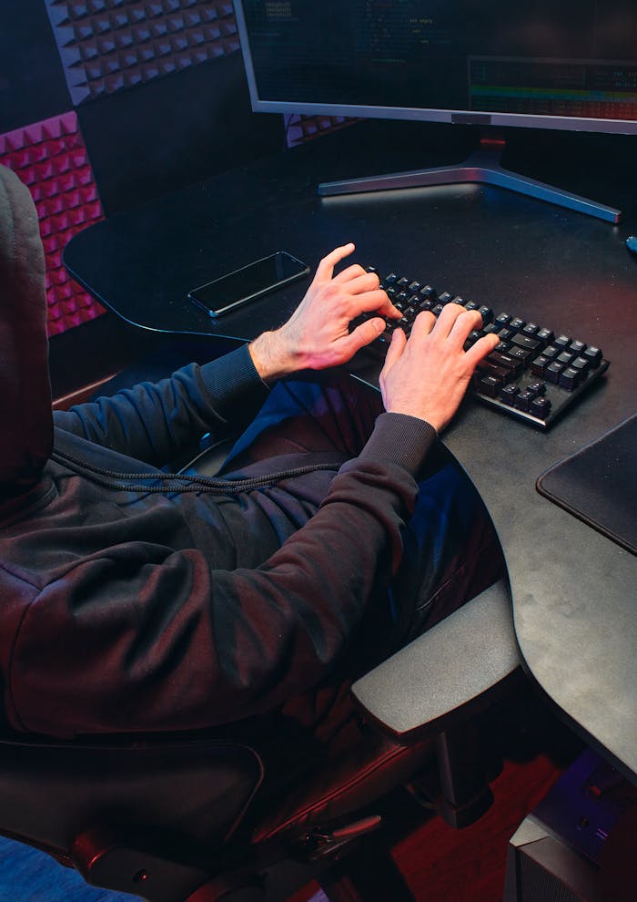 Person in hoodie typing on a keyboard in a dark room, focusing on hacking activities.